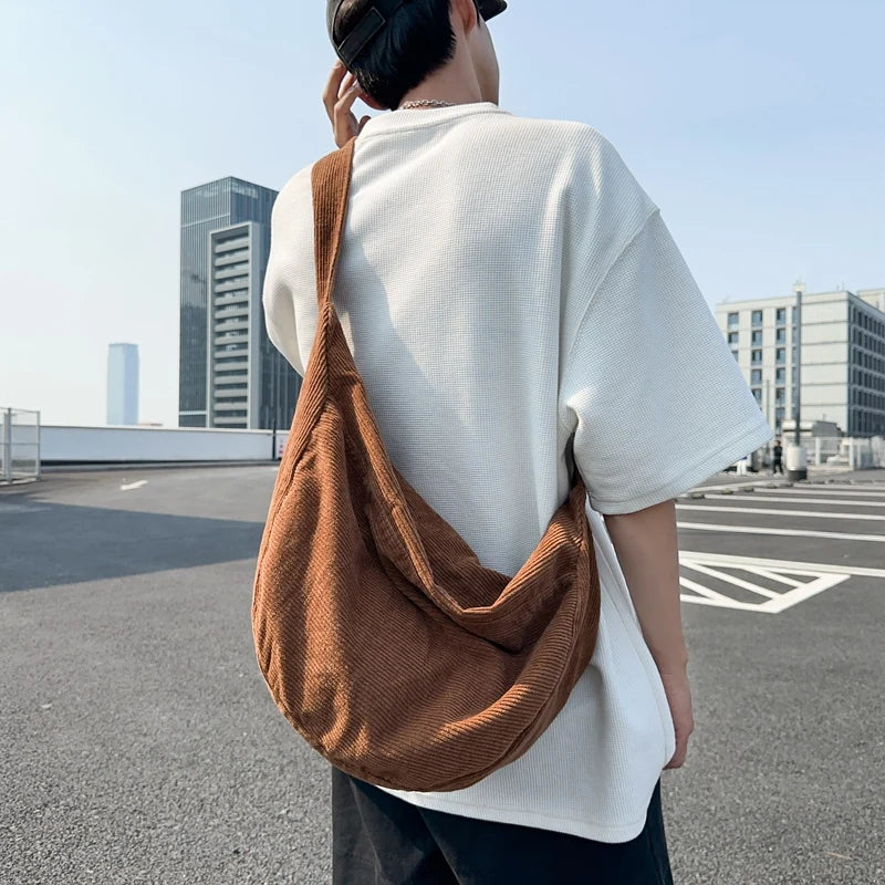 A person wearing a white t-shirt and dark pants is holding a brown corduroy casual crossbody shoulder bag.