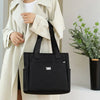 A black nylon shoulder tote bag is held by a person wearing a beige trench coat.