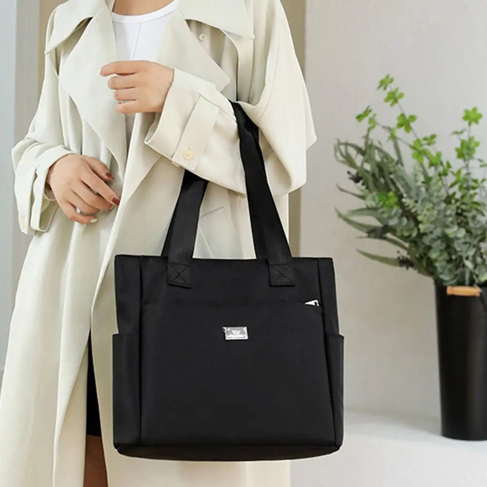 A black nylon shoulder tote bag is held by a person wearing a beige trench coat.