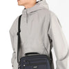 A man wearing a light gray waterproof jacket is holding a navy blue compact crossbody messenger bag.