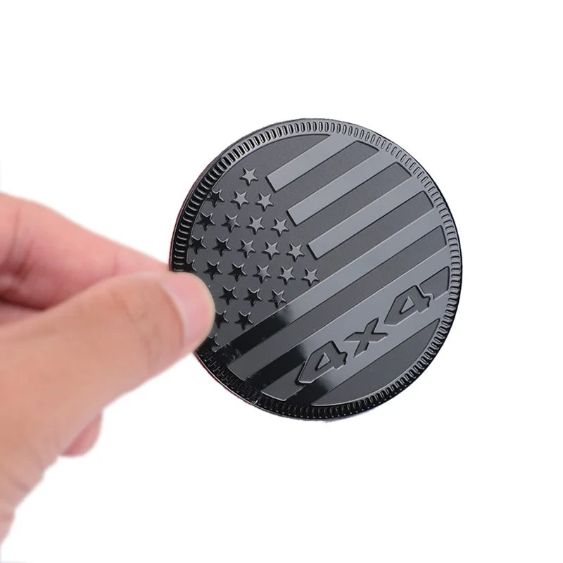 A hand holds a round black aluminum alloy car decal featuring the American flag design and 4x4 text.