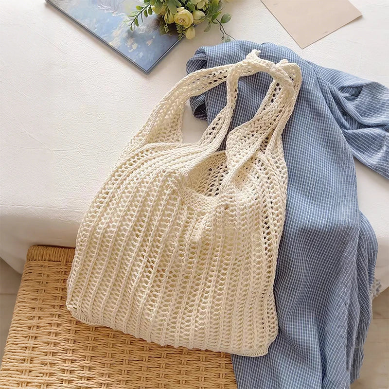 A cream-colored bohemian knit shoulder bag resting on a woven mat next to a blue plaid fabric.