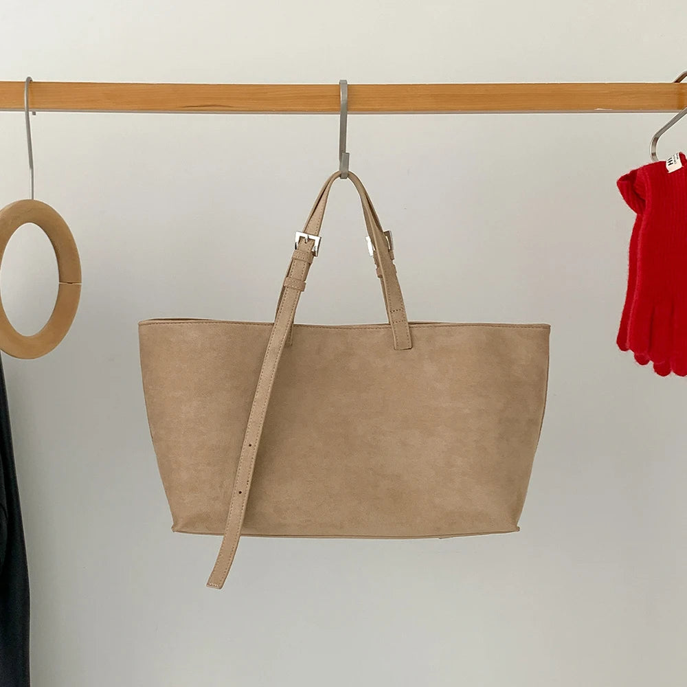 Un sac à main en cuir PU beige et décontracté est suspendu sur un support en bois contre un mur blanc.