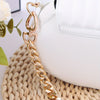 A close-up of a white leather purse with a gold heart-shaped metal chain extender resting on a woven surface.