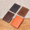 Three genuine leather bifold card holders in dark brown, reddish-brown, and orange colors arranged on a wooden surface.