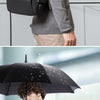 A man wearing a gray shirt and suit jacket carries a black business travel backpack under an umbrella.