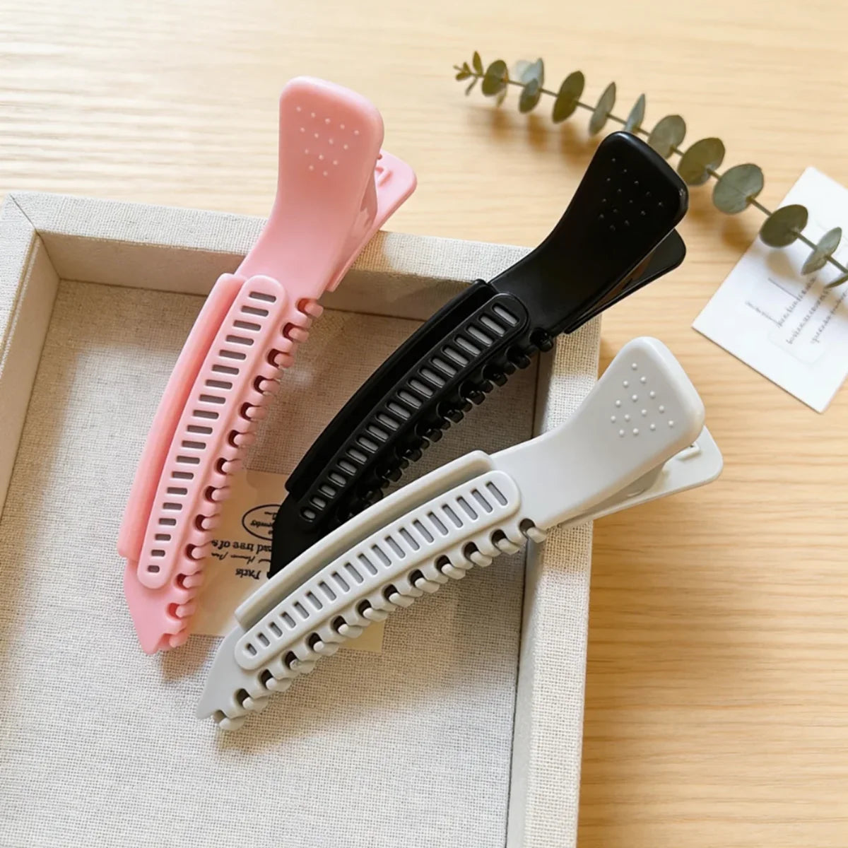 Trois pinces à cheveux professionnelles antidérapantes roses et noires dans une boîte en bois sur une surface en bois clair.