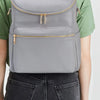 A woman wearing a green shirt and dark jeans is shown holding a gray large capacity diaper bag backpack.
