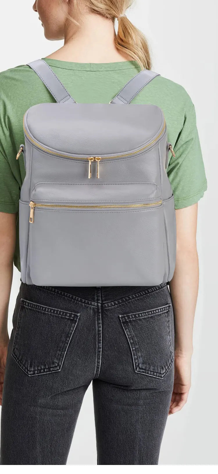 A woman wearing a green shirt and dark jeans is shown holding a gray large capacity diaper bag backpack.