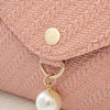A close-up of a dusty rose woven texture fabric on a crossbody bag featuring a gold-toned button and a pearl charm.