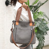 A woman holds a gray canvas tote bag with brown leather straps against a white wall, next to a large green plant.