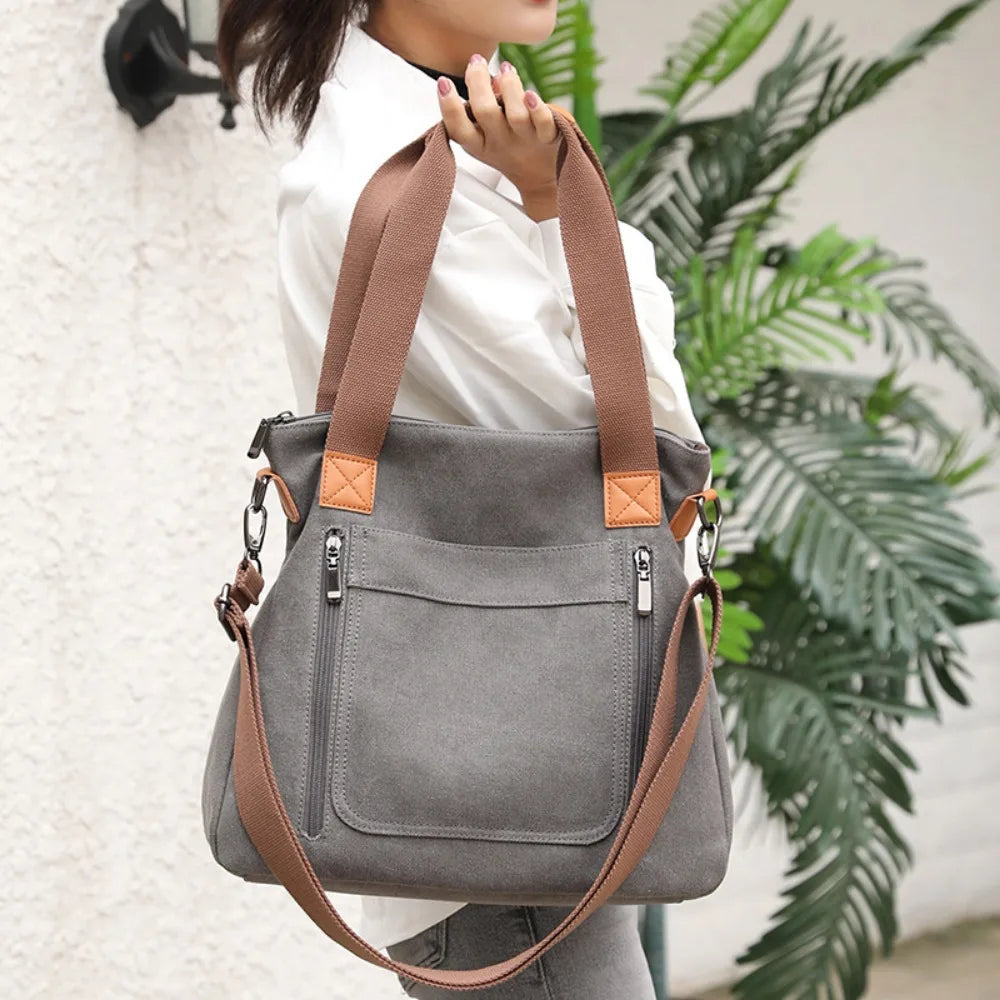 A woman holds a gray canvas tote bag with brown leather straps against a white wall, next to a large green plant.