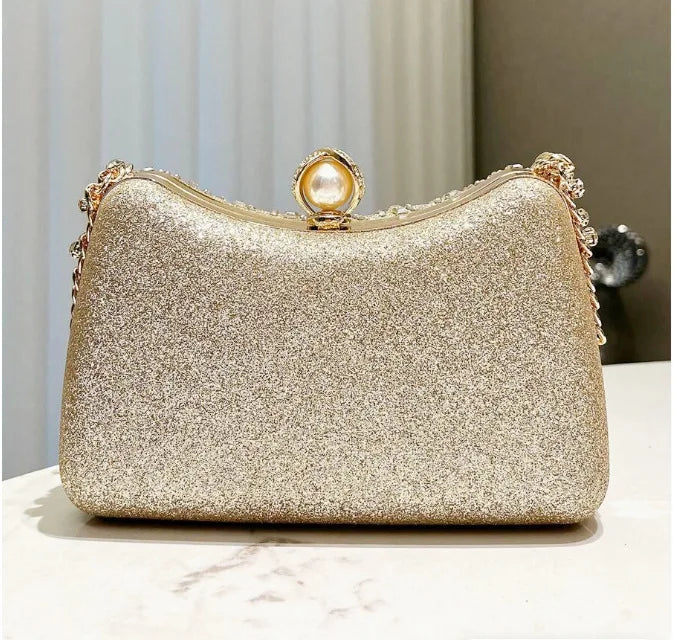 A gold glitter clutch bag with a pearl embellishment and rhinestone detailing resting on a white surface.