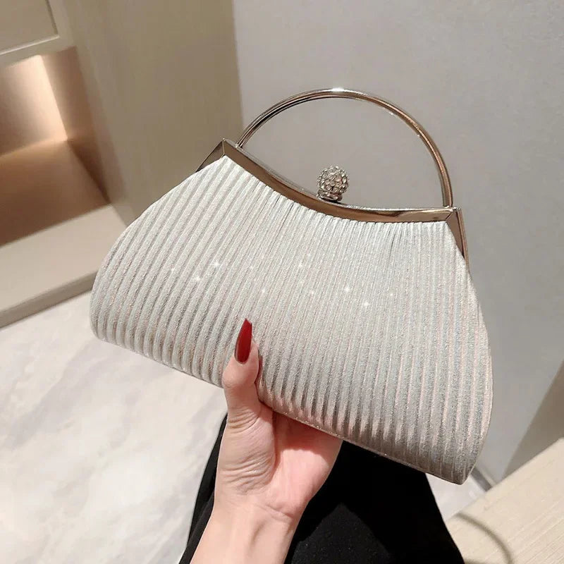 A light beige pleated glitter evening handbag with a gold top handle and chain strap being held by a hand.