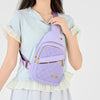 Une femme tient un sac bandoulière matelassé violet avec des fermetures éclair dorées contre une chemise bleu clair.