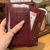 A hand holds a maroon leather passport wallet with RFID blocking features and a card slot.