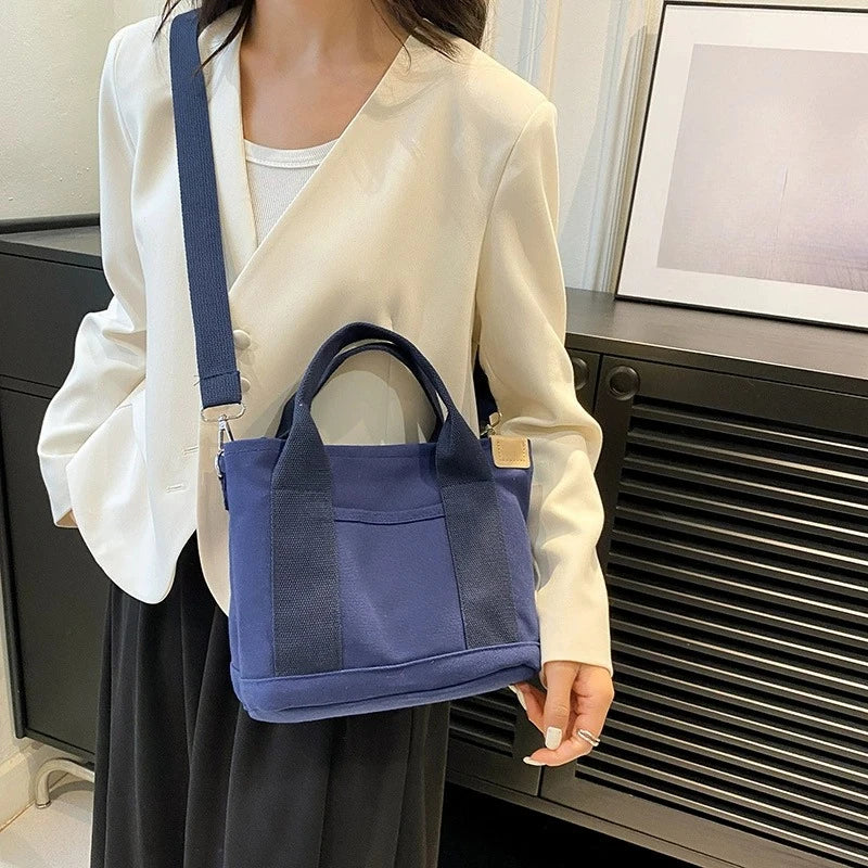 A woman wearing a cream-colored blouse and dark skirt holds a navy blue canvas crossbody bag.