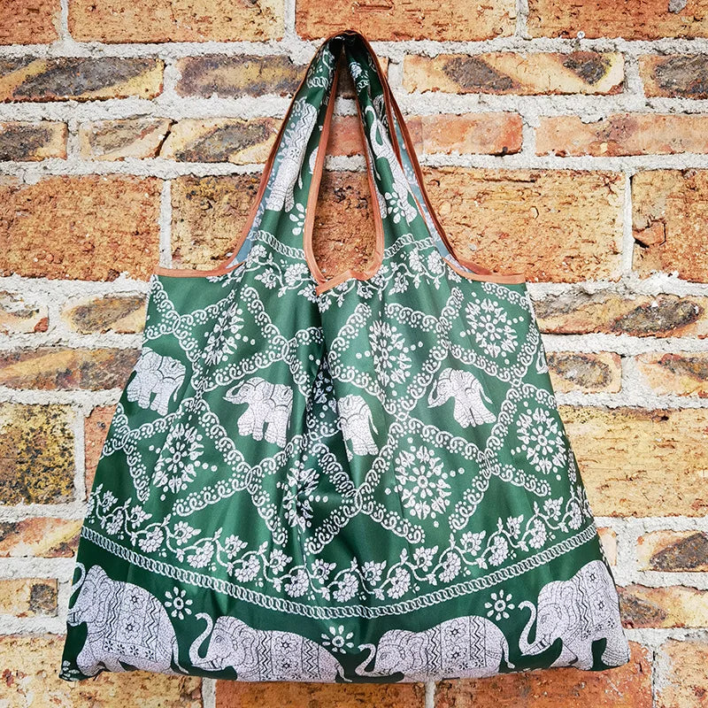 A reusable shopping bag featuring a green and white patterned fabric with elephant motifs, displayed against a brick wall.