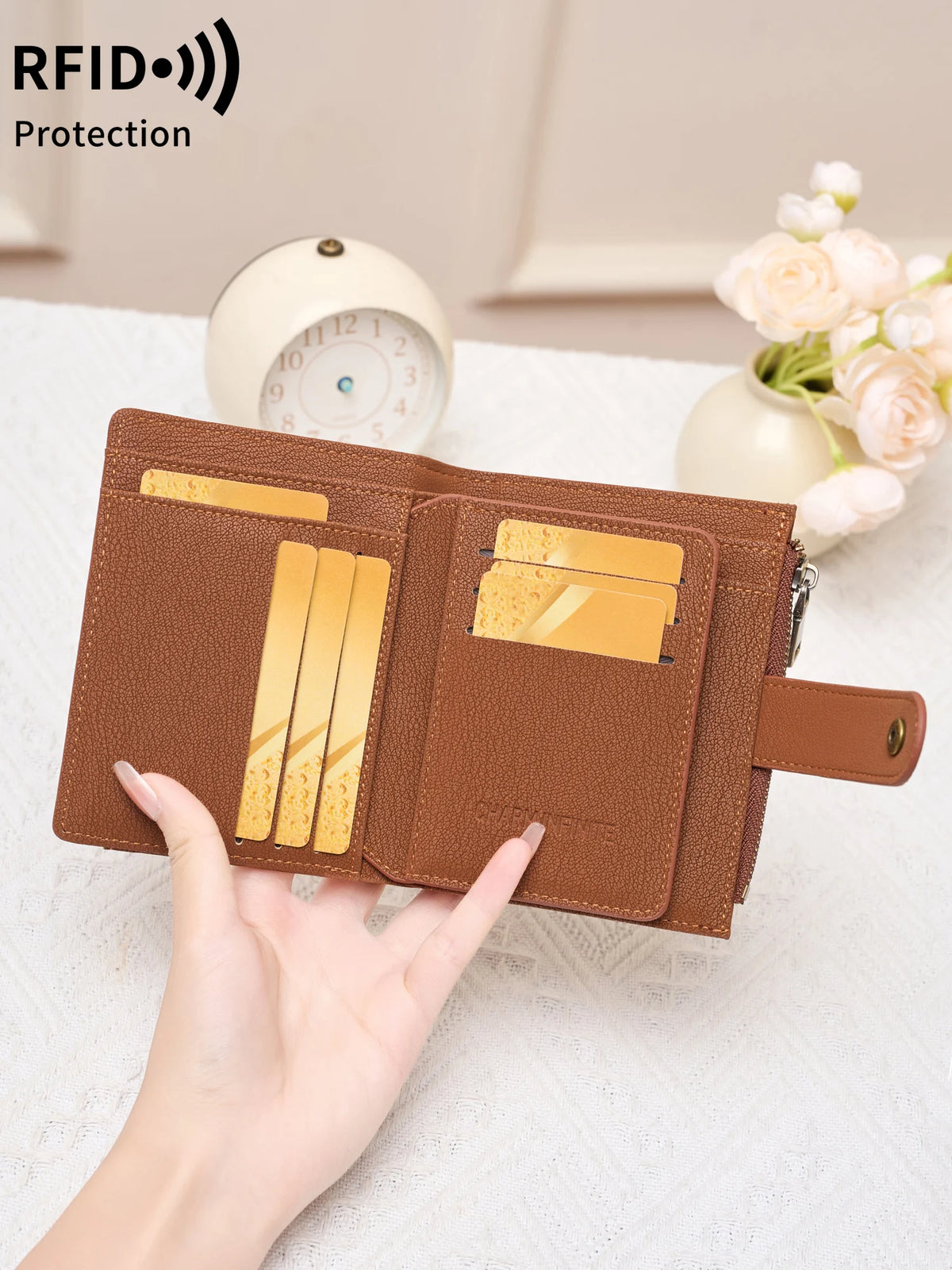 A hand holds a brown RFID-blocking compact bifold zipper wallet with gold cards and a white alarm clock in the background.