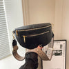 A black PU leather crossbody bag with a gold-toned adjustable strap is displayed against a light wall.