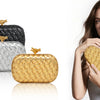 A woman holds a gold woven clutch bag next to silver and black versions.