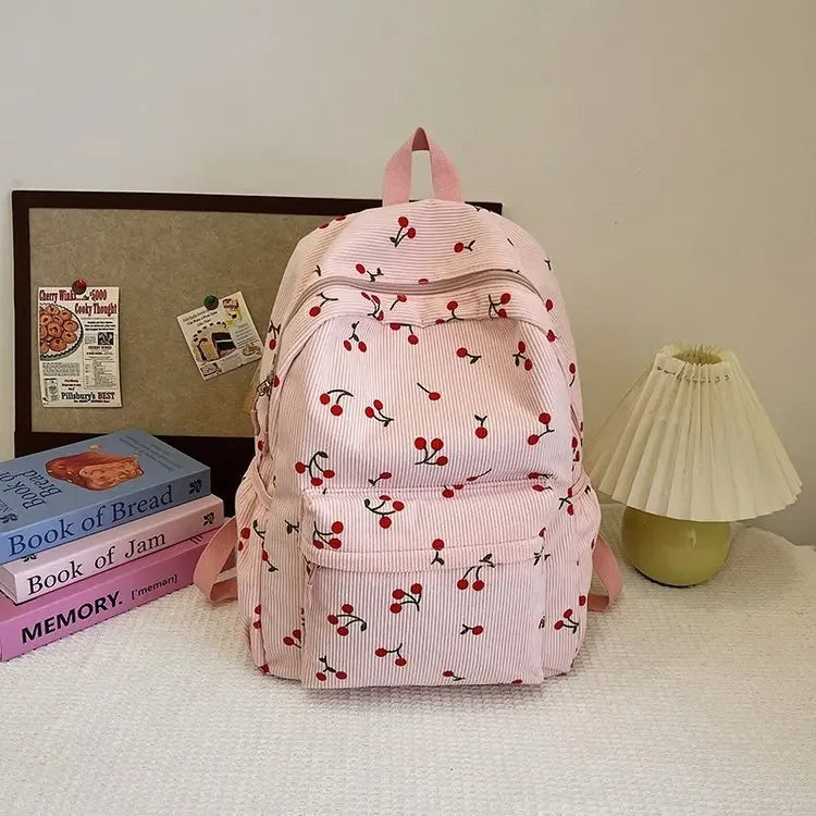 A pink corduroy fashion backpack with a red cherry pattern resting on a white surface.