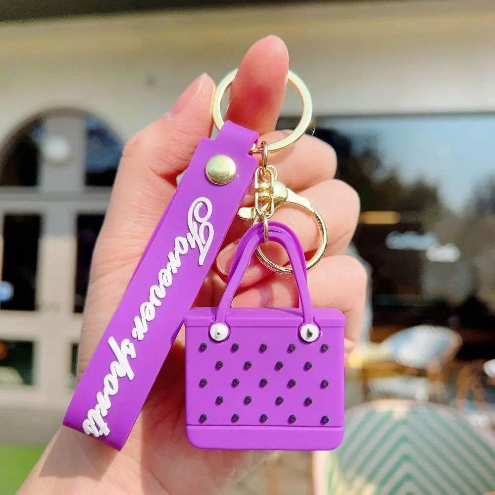 A purple silicone heart beach bag keychain is held in a hand against a blurred outdoor background.