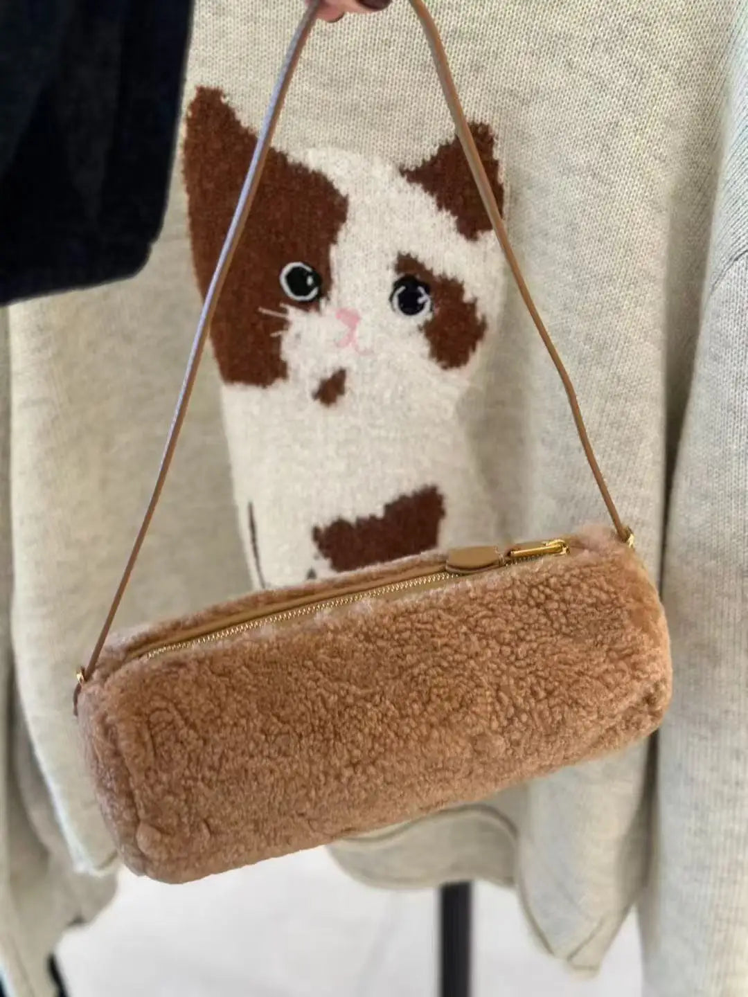 A small brown faux fur cylinder shoulder bag with a gold zipper is being held by a cat.