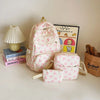A pink nylon travel backpack set with a bow print is displayed next to a lamp and books.