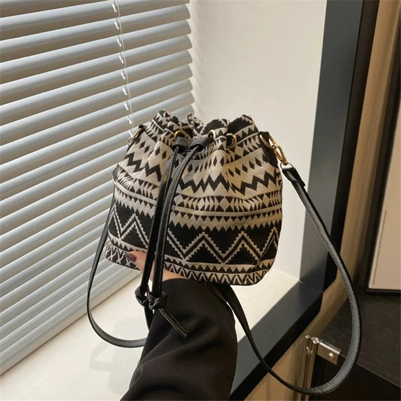 A close-up of a patterned black and beige floral drawstring bucket bag being held by a person near a window blind.