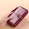 A hand holds a maroon wax leather long wallet with a wrist strap, showing the texture and design.