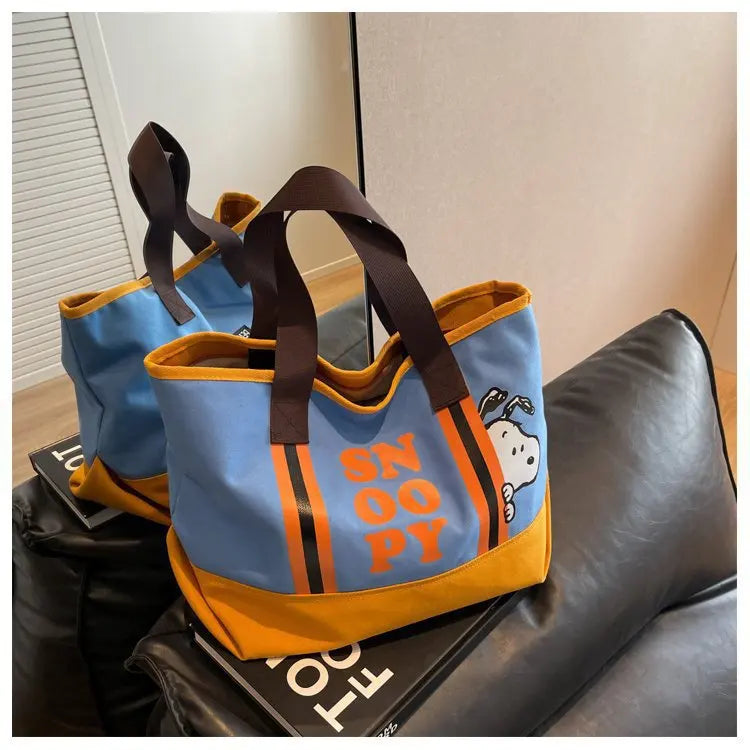 A blue and orange canvas shoulder bag featuring a Snoopy cartoon design resting on a dark leather couch.