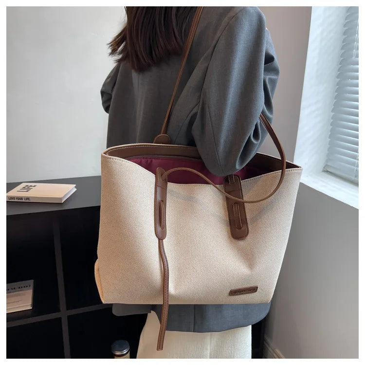 A light beige canvas shoulder tote bag with brown leather accents is displayed next to a dark surface.