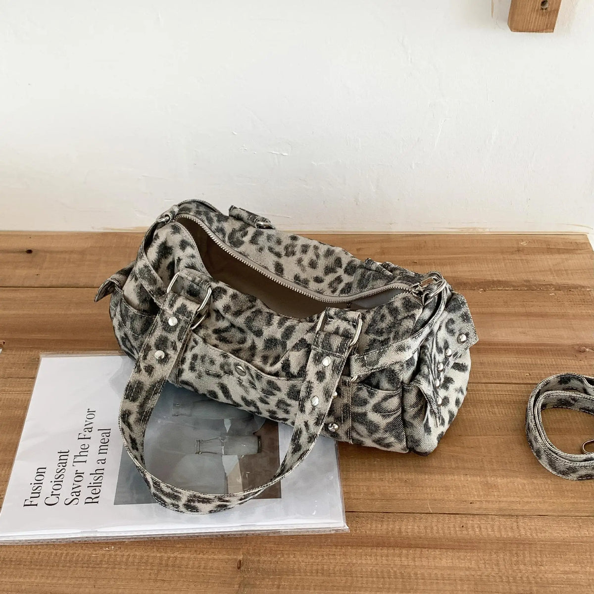 A leopard print riveted Boston shoulder bag resting on a wooden surface next to a magazine.