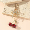 Pendentif de cerise rouge doré avec nœud scintillant, posé sur une surface beige texturée.