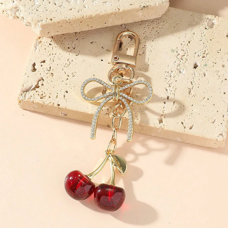 Pendentif de cerise rouge doré avec nœud scintillant, posé sur une surface beige texturée.
