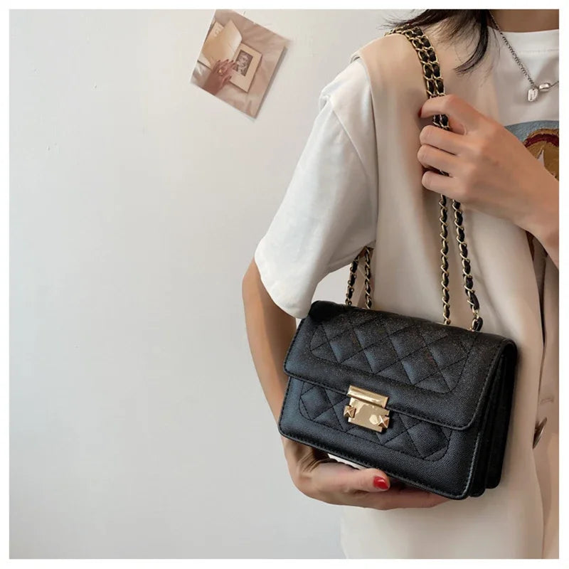 A hand holds a black quilted crossbody bag with a gold clasp and a metal shoulder strap against a white shirt.