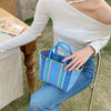 A woman wearing a white top sits outdoors holding a blue and green hand woven PVC vegetable basket tote bag.