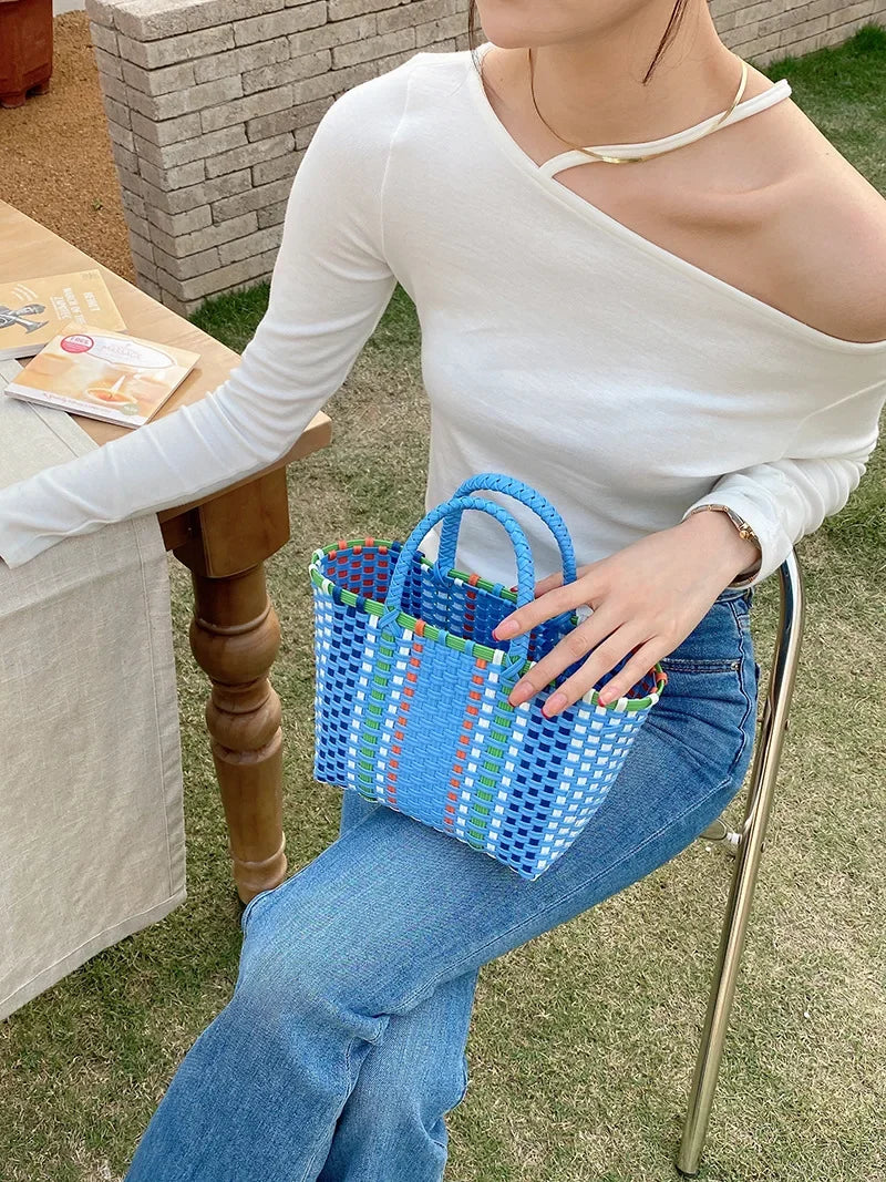 A woman wearing a white top sits outdoors holding a blue and green hand woven PVC vegetable basket tote bag.