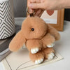 A fluffy, light brown plush rabbit charm with a gold keyring is being held by a hand.