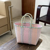 A hand woven pink and yellow striped PVC vegetable basket tote bag sitting on a dark wooden surface.