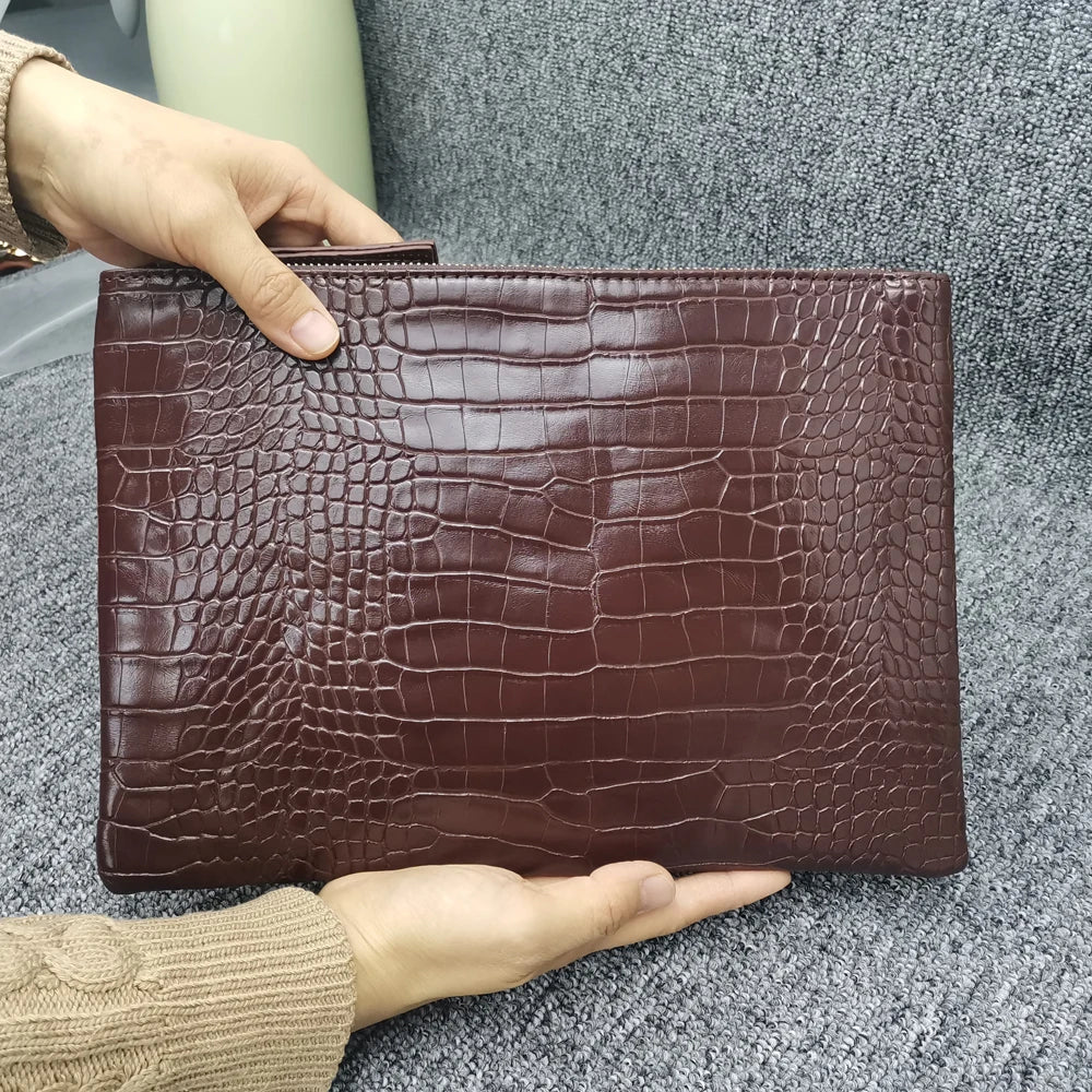 A hand holds a brown alligator print clutch bag against a gray textured background.