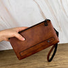 Une main tient un portefeuille/sacoche retro en cuir marron pour homme sur un fond de marbre clair.