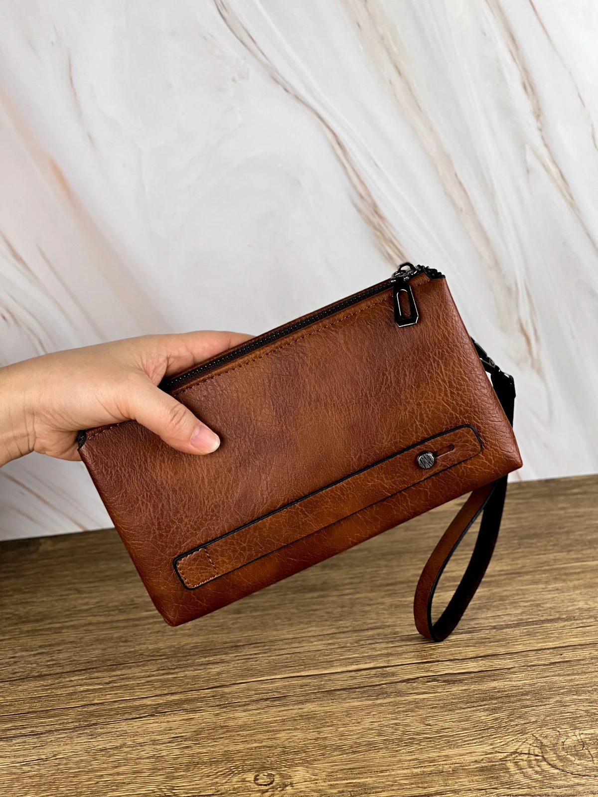 Une main tient un portefeuille/sacoche retro en cuir marron pour homme sur un fond de marbre clair.