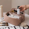 A hand holds a beige foldable cosmetic storage bag with a lid open, showing various beauty products and brushes.