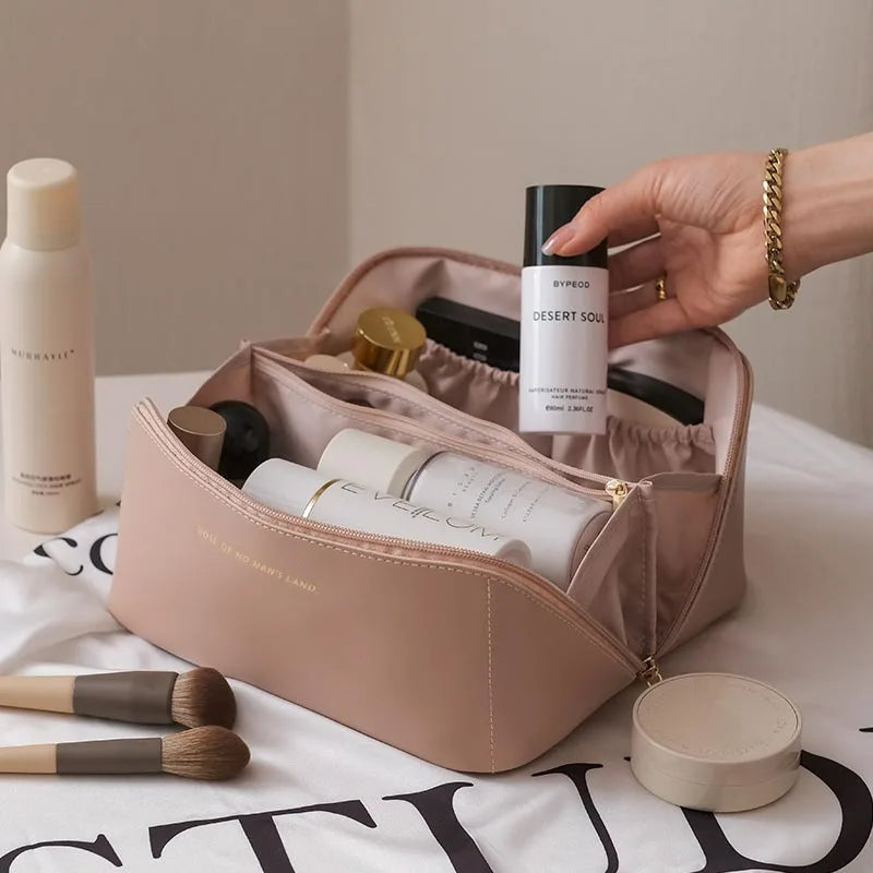A hand holds a beige foldable cosmetic storage bag with a lid open, showing various beauty products and brushes.