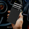 A hand holds a black carbon fiber magnetic card holder wallet with a card inserted, set against a car interior background.