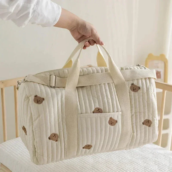 Una bolsa de pañales acolchada beige con parches de oso marrón claro es sostenida por una mano.