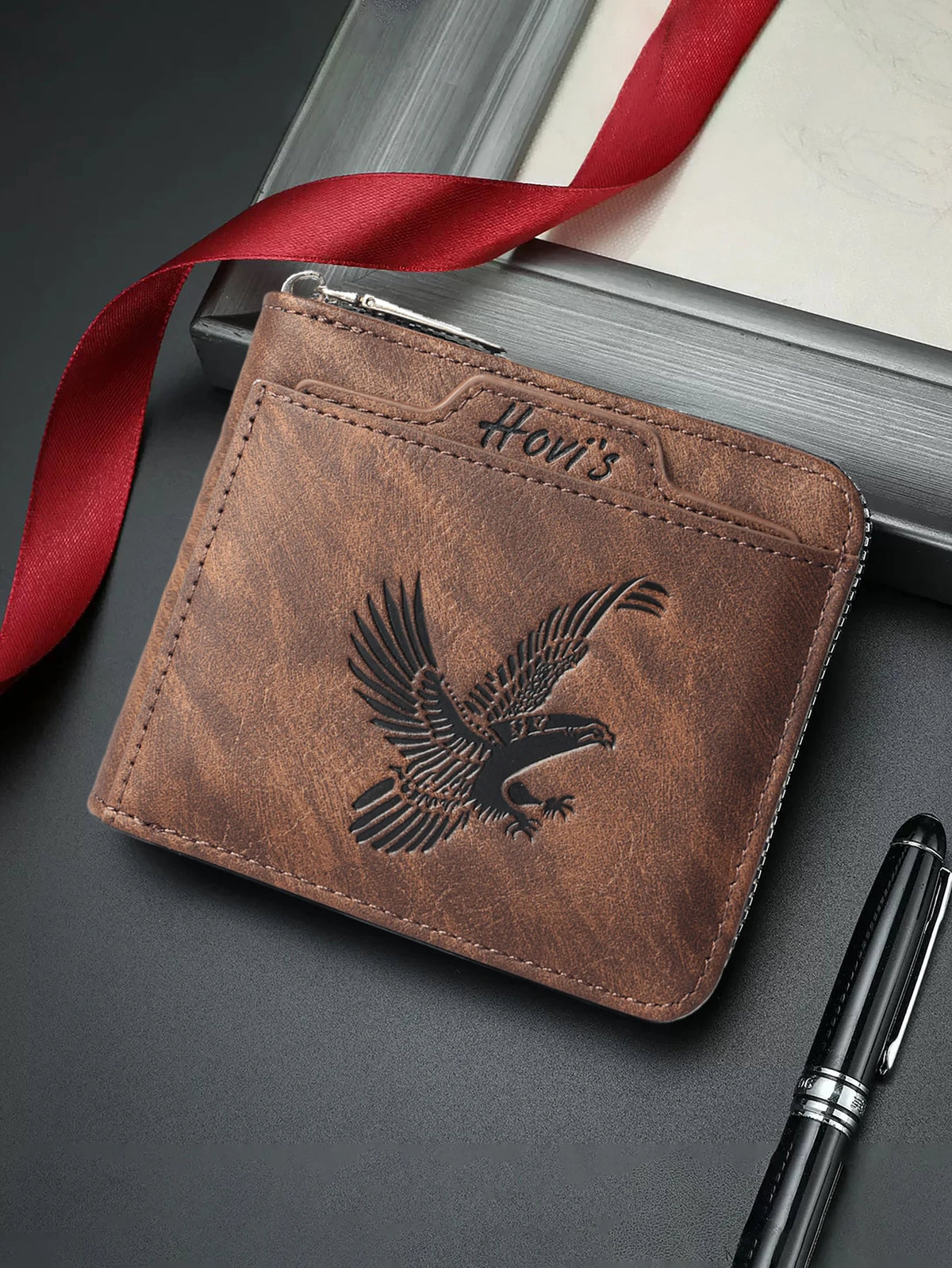 A brown embossed PU leather slim wallet with a silver clasp and a red ribbon, resting on a dark surface next to a black pen.