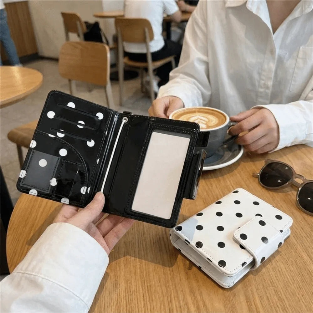 A black polka dot passport holder is shown on a wooden table next to a white polka dot wallet.